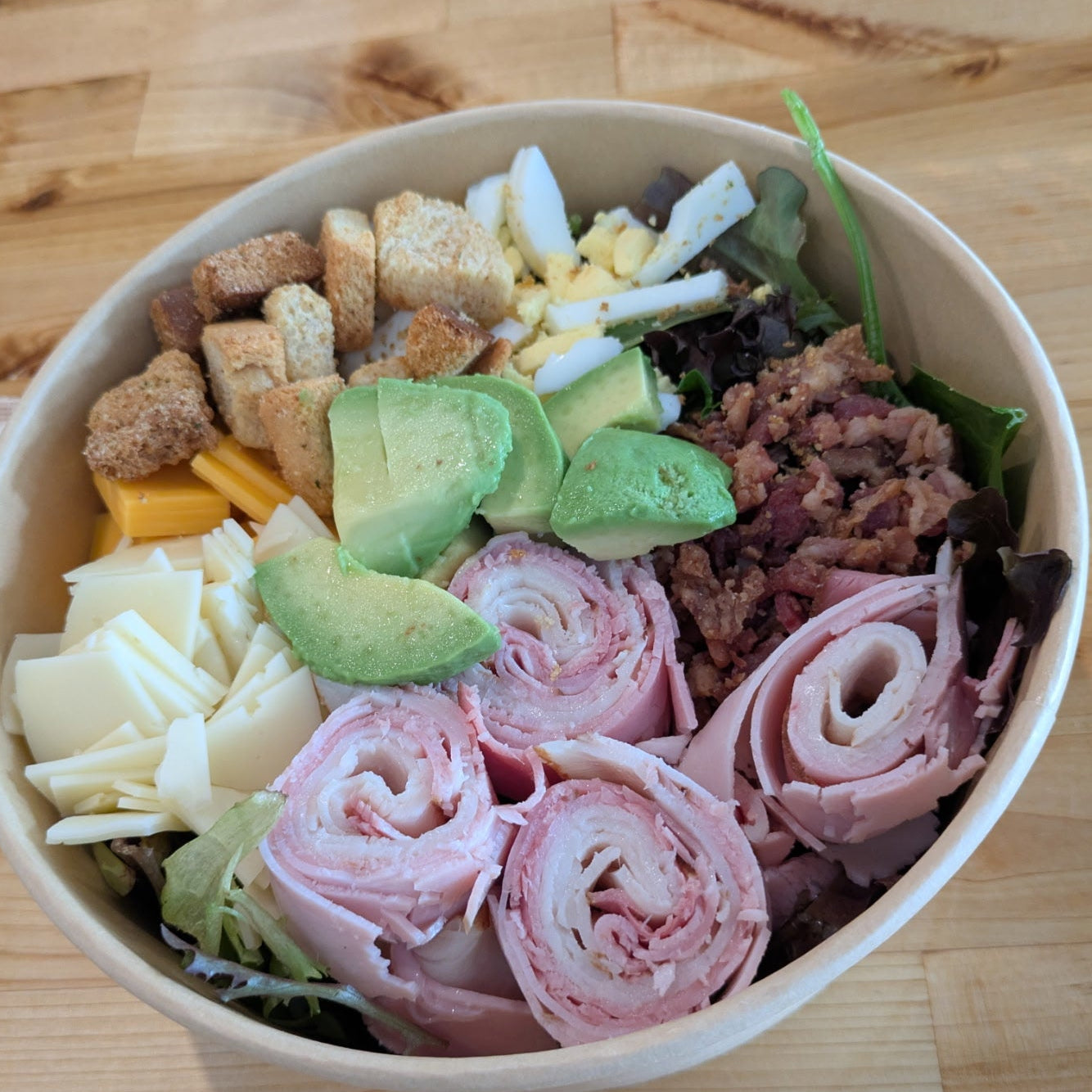 Salad bowl with various ingredients including ham, cheese, and avocado on a wooden surface