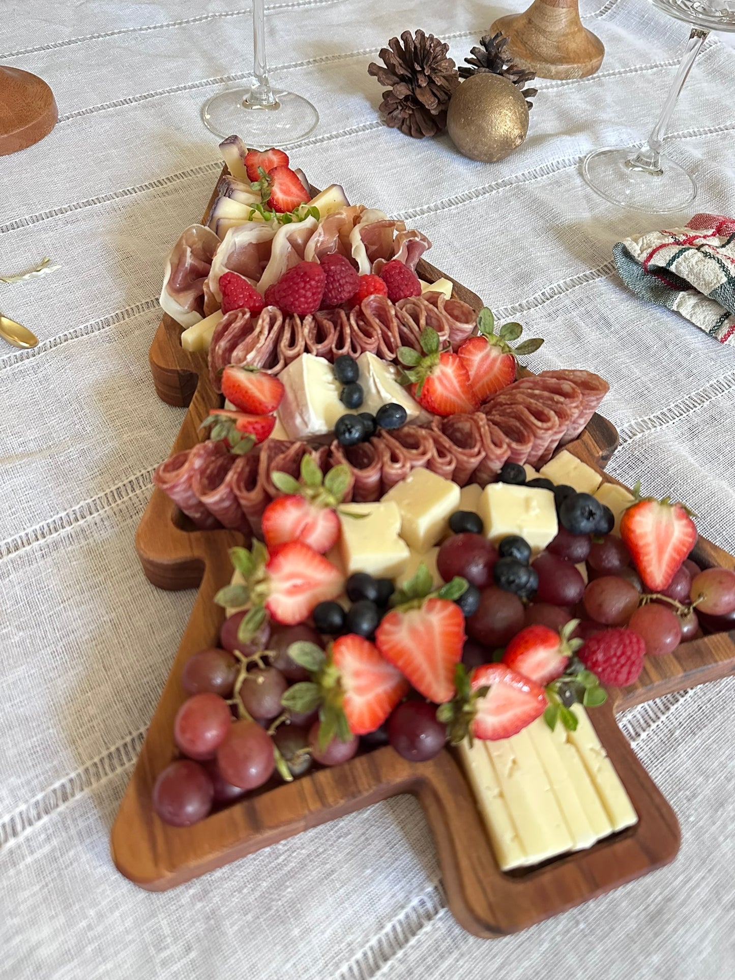 Wooden Christmas tree-shaped charcuterie board with fruits and meats on a white tablecloth.