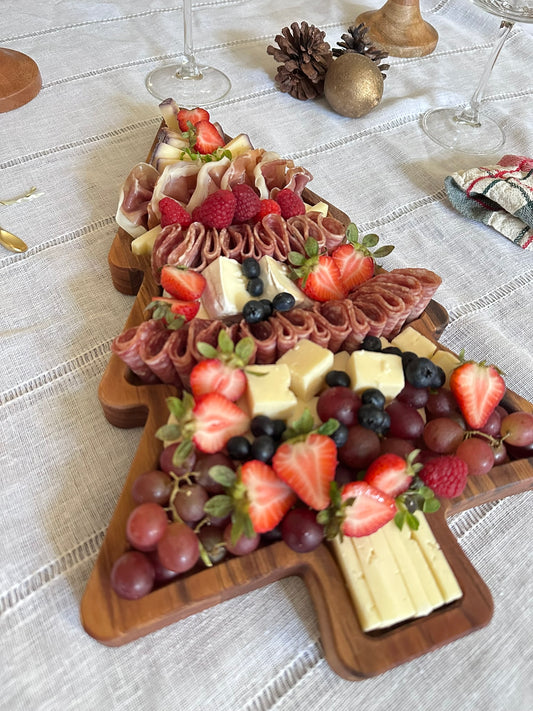 Wooden Christmas tree-shaped charcuterie board with fruits and meats on a white tablecloth.