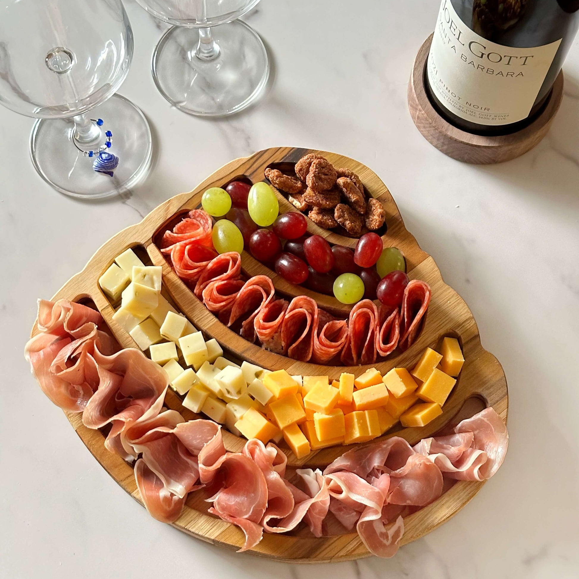 Wooden charcuterie board shaped as the Pawleys Island shell, with meats, cheeses, and fruits on a marble surface with wine glasses and a bottle.