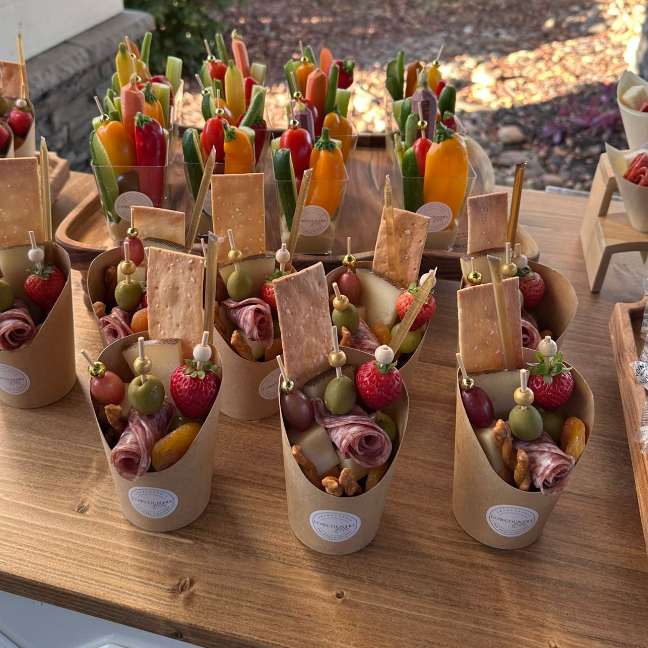 Assorted charcuterie, fruit and vegetable skewers in paper cups on a wooden table outdoors.