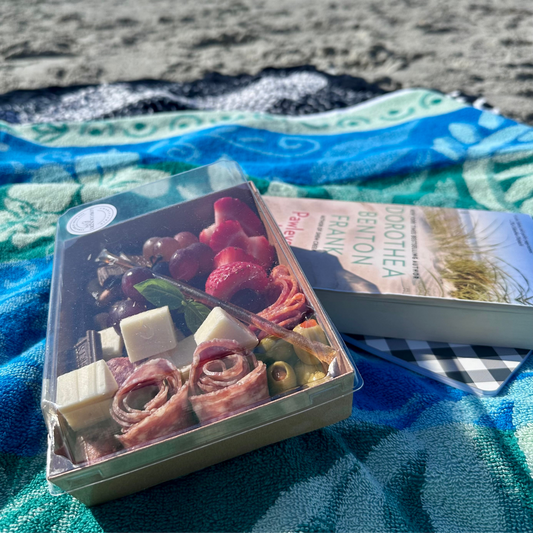Box of assorted meats and cheeses on a beach with a magazine underneath
