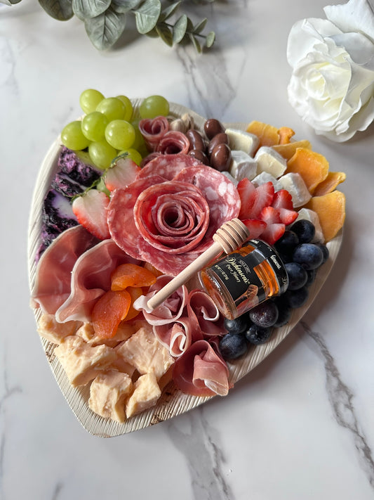 A heart-shaped charcuterie board arranged with an assortment of artisan cheeses, cured meats, fresh garnishes, and chocolates, presented on a white surface.