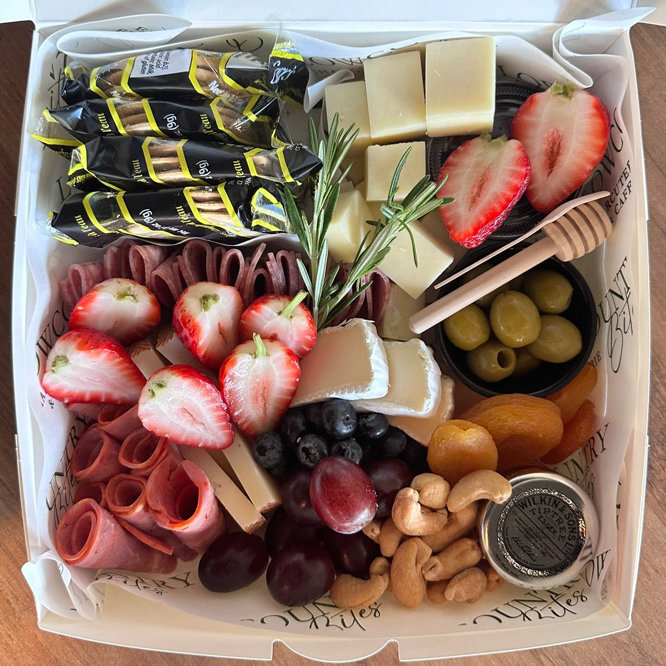 Assorted charcuterie board with fruits, meats, and cheeses in a white box on a wooden surface.